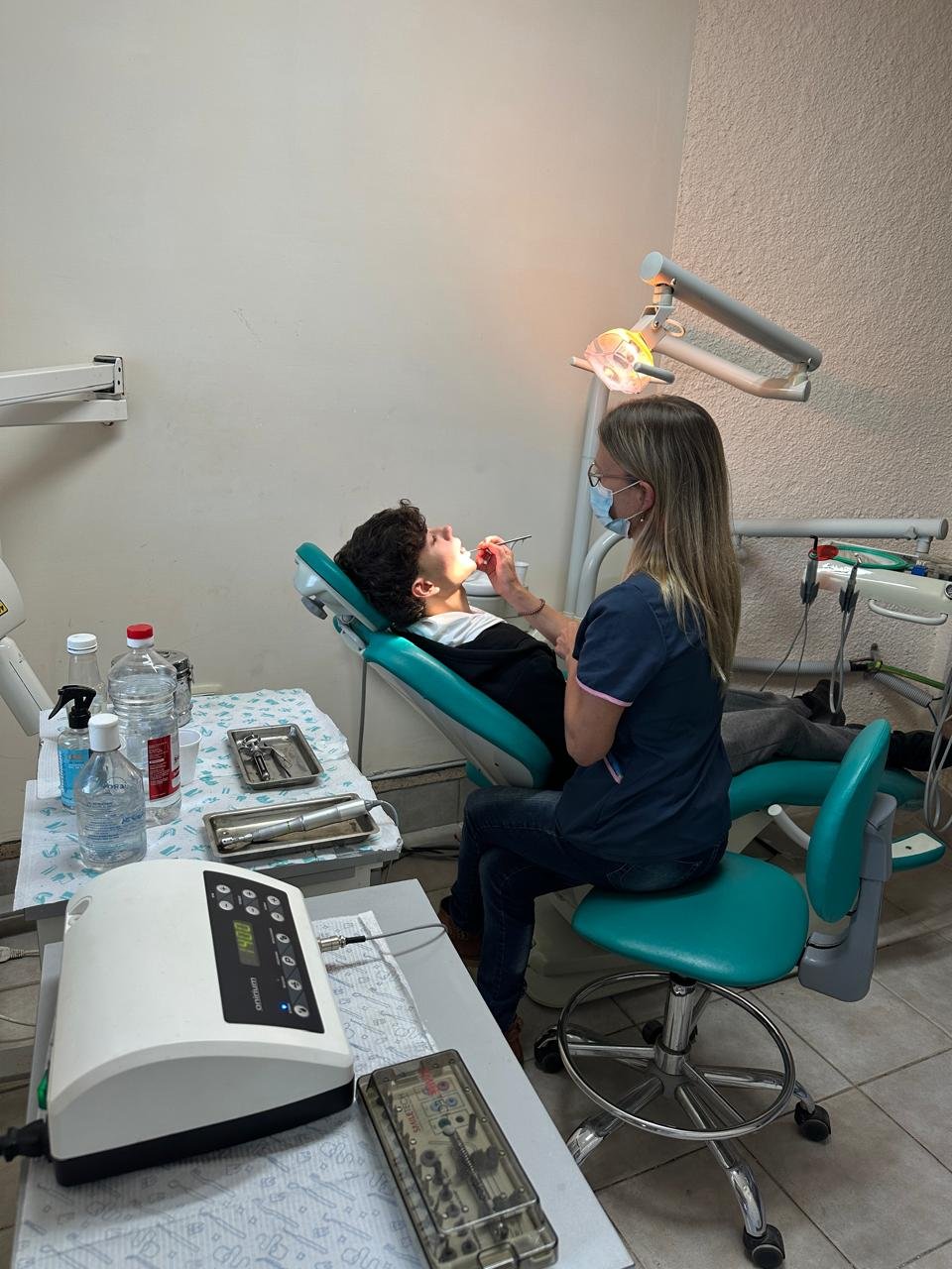Dentist in blue mask and scrubs examining a patient in a teal dental chair under overhead light