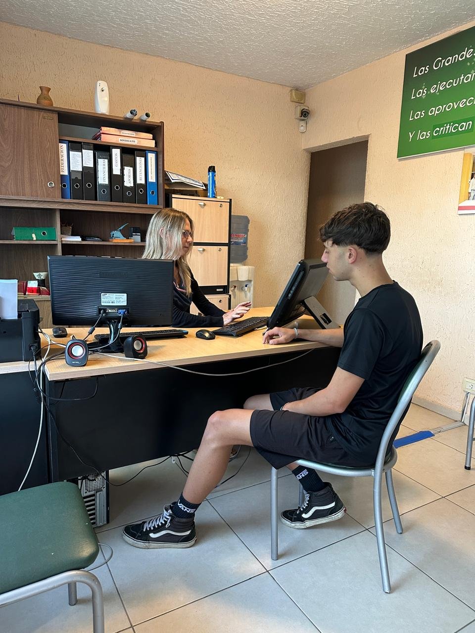 Two young people sitting at a desk with a computer in a classroom setting with educational posters on the wall