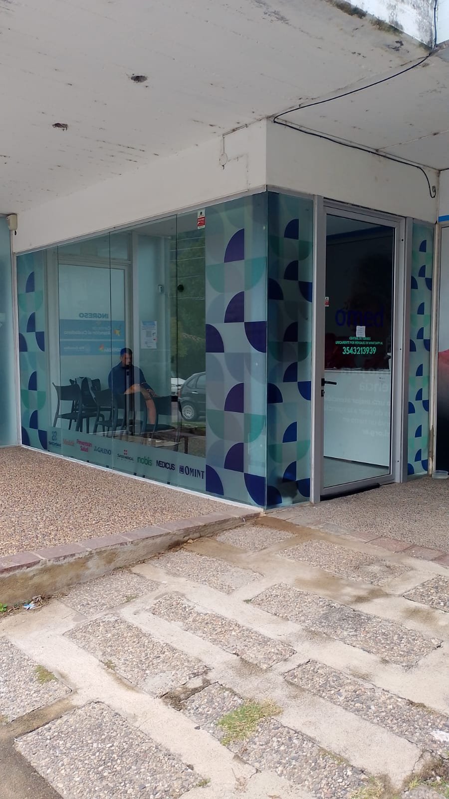 Small modern storefront with turquoise and navy geometric pattern on glass frontage, white ceiling, and concrete paver walkway