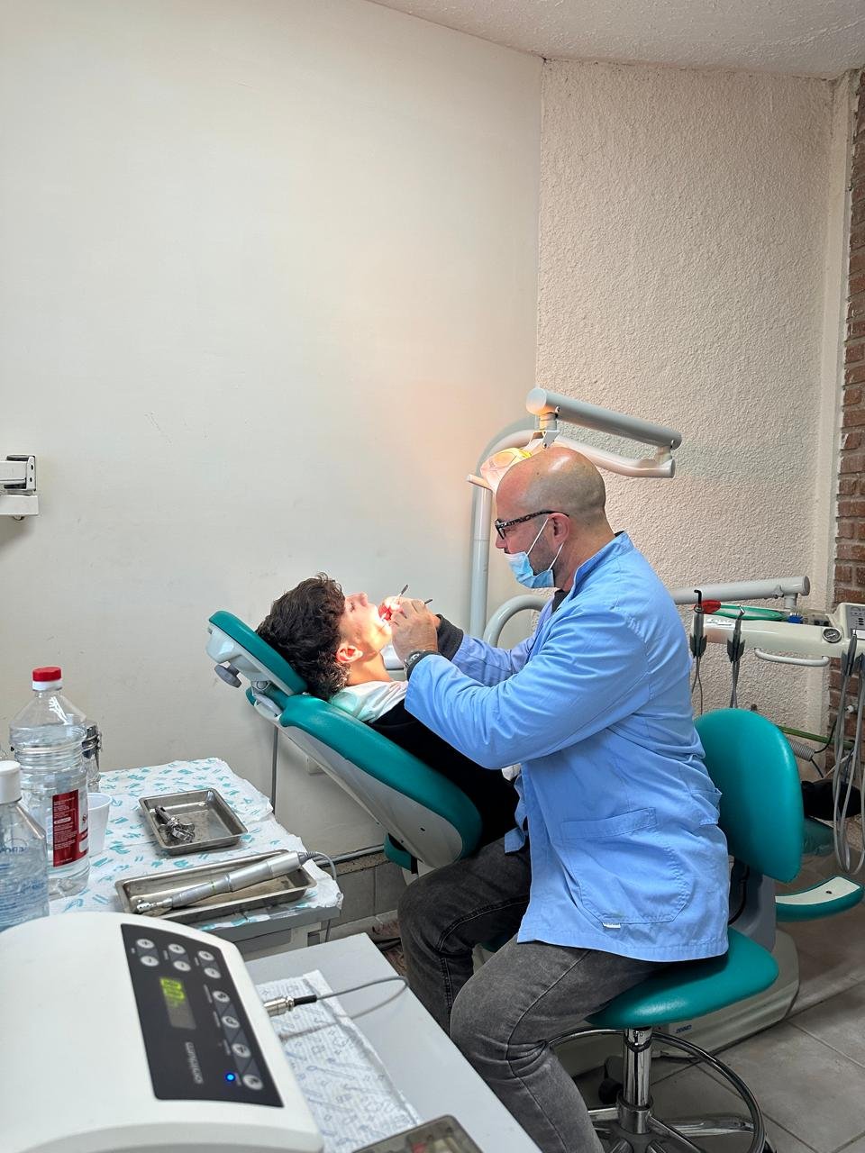Dentist examining patient in dental chair with overhead light and equipment visible in clinic